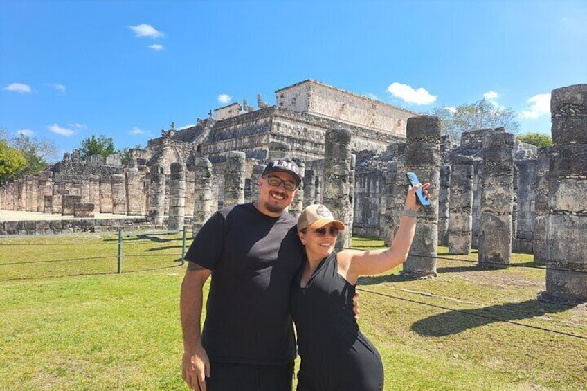 Two people taking a selfie in front of ancient ruins with stone columns and a large building in the background. 1