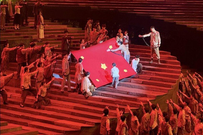 A group of people on a stage with a red backdrop, some standing and others sitting, in a theatrical setting. 1