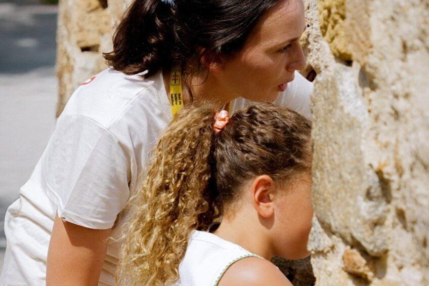 A person with curly hair tied back, wearing a white shirt, standing next to a stone wall. 3