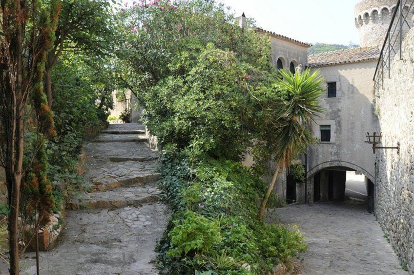 A stone pathway leading through a lush garden, with a building featuring arched windows and a tower in the background. 5