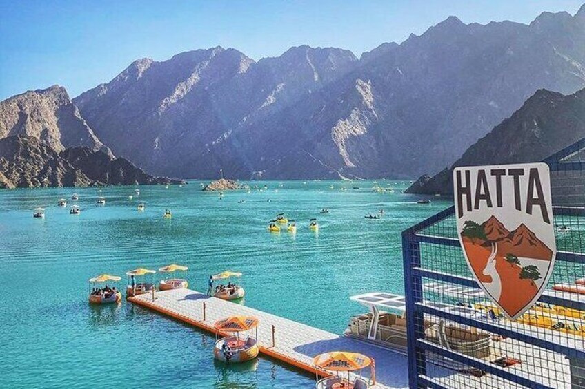 Escena costera con un letrero que dice "HATTA" y un muelle con barcos y sombrillas. 2