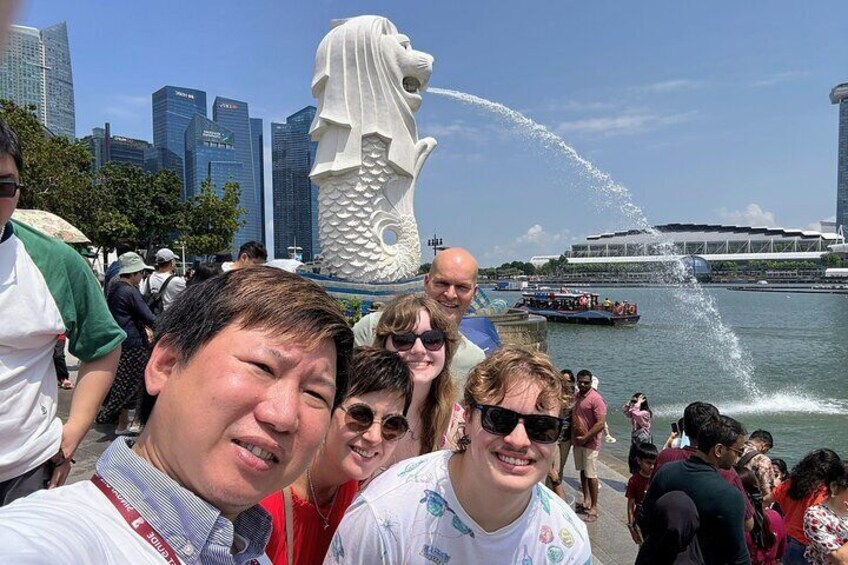 Un gruppo di persone vicino a una grande statua bianca con getti d'acqua, con sullo sfondo uno skyline cittadino e una nave da crociera. 1