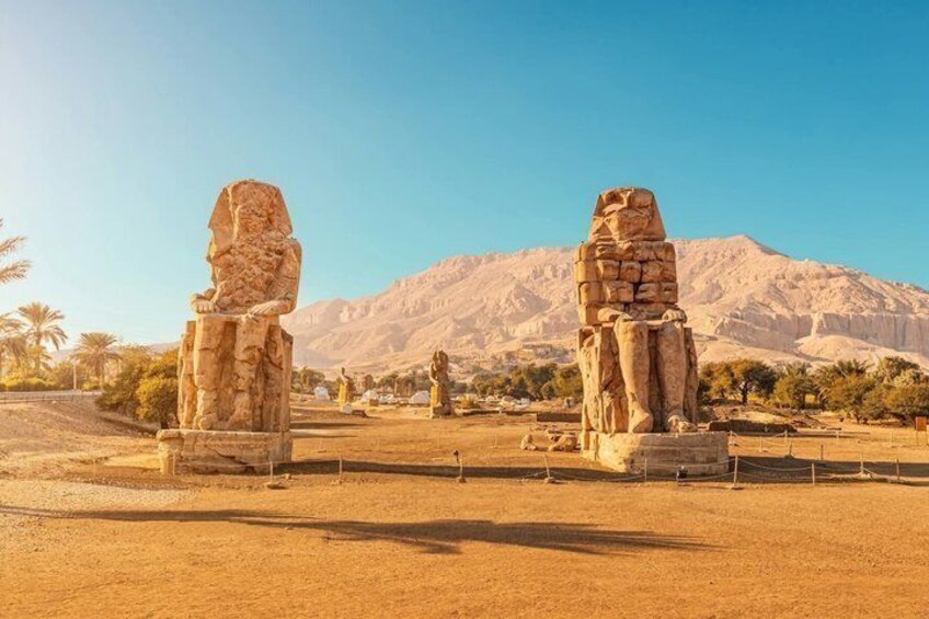 Deux grandes statues de pierre se dressent dans un paysage désertique, avec des montagnes en arrière-plan. 1