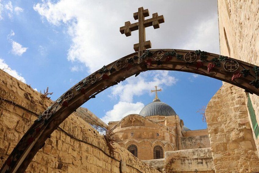 A stone archway with a cross, leading to a building with a dome and a cross on top. 3