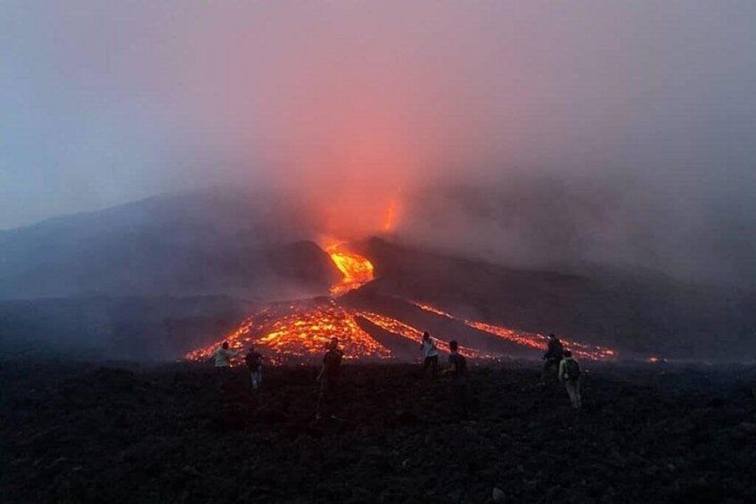 A volcano with lava flowing down its side, surrounded by a group of people observing. 1