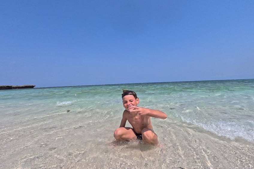 A person sitting on a sandy beach with clear turquoise water and a clear sky. 3