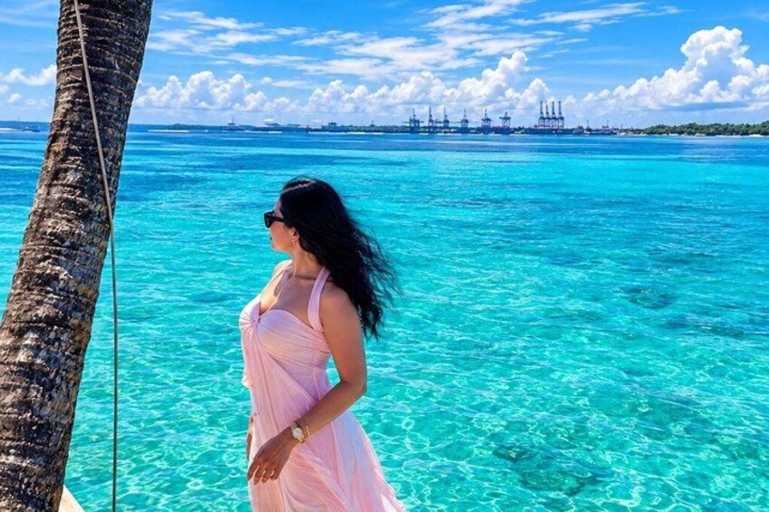 A woman in a pink dress standing by a palm tree, looking at the turquoise ocean and distant industrial structures. 2