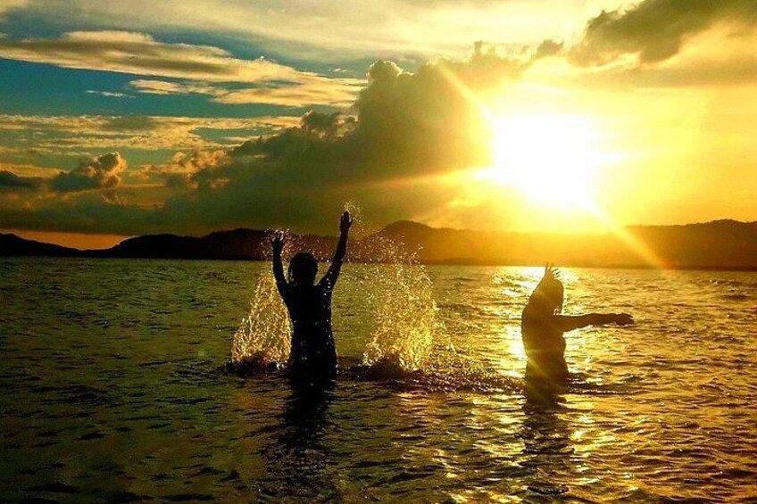 Eine Person im Wasser mit erhobenen Armen, eine weitere Person im Hintergrund und ein Sonnenuntergang über dem Wasser. 3