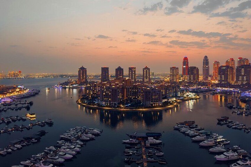 A city skyline at dusk with illuminated buildings, a marina filled with boats, and a calm waterfront. 2