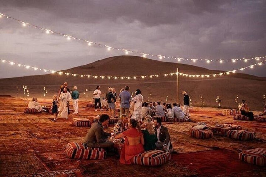 A group of people sitting on cushions in a desert setting with string lights overhead. 1