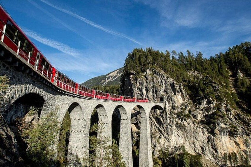 A red train on a stone viaduct with multiple arches, surrounded by forested mountains. 4
