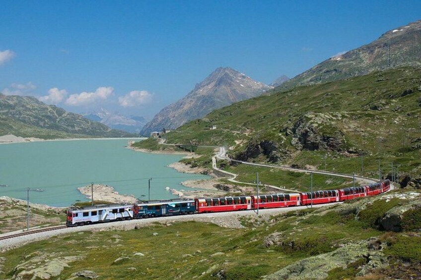 A red train on a mountain track with a lake and mountains in the background. 2