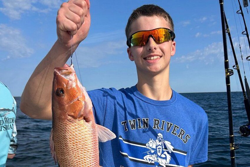 A person holding a fish, wearing a blue shirt with 'Twin Rivers' text, standing on a boat with fishing rods in the background. 4
