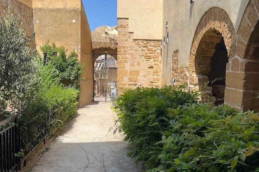 Un estrecho callejón con paredes de piedra, una reja metálica y abundante vegetación. 4
