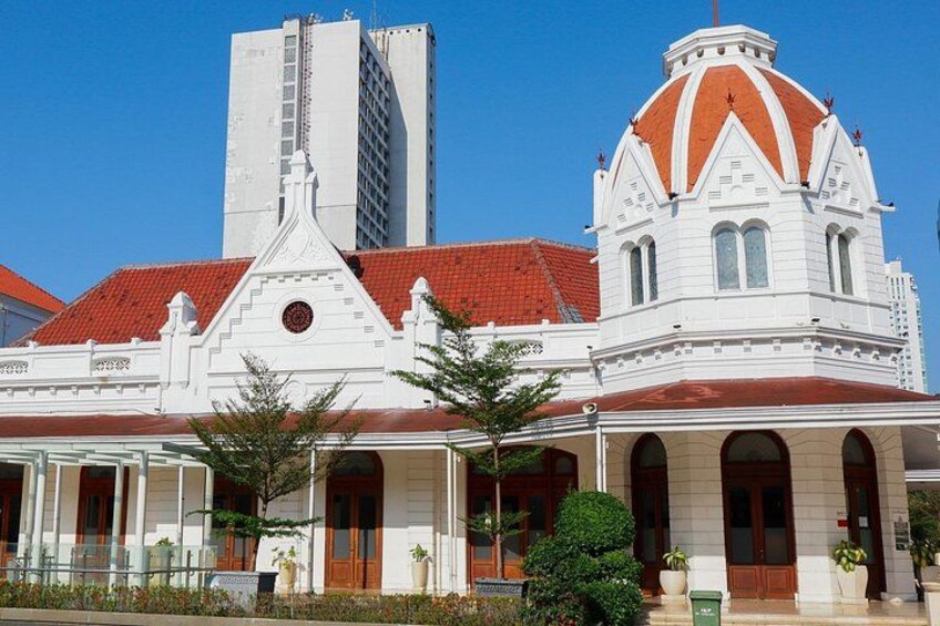 A white building with a red roof and a dome, surrounded by greenery and a tall building in the background. 2