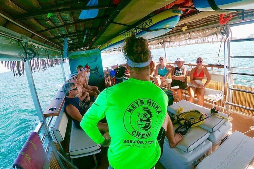 Un barco con gente en la cubierta, una persona con camisa verde con un logotipo, y un paisaje tropical. 5