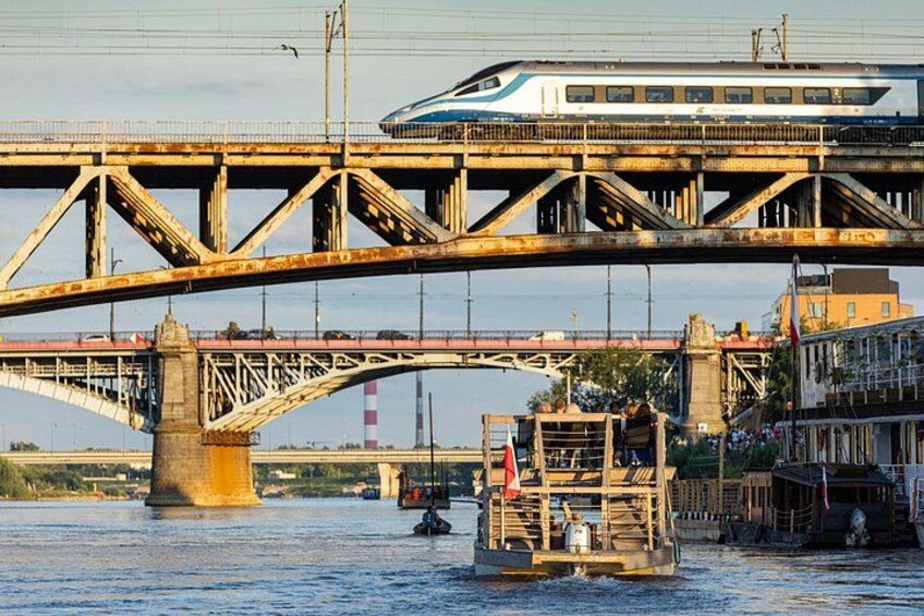 A train crossing a bridge over water, with a boat in the foreground and buildings along the riverbank. 5