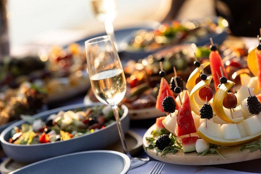 A table with a variety of food items, including skewers with fruits and nuts, a glass of white wine, and plates of salad. 2