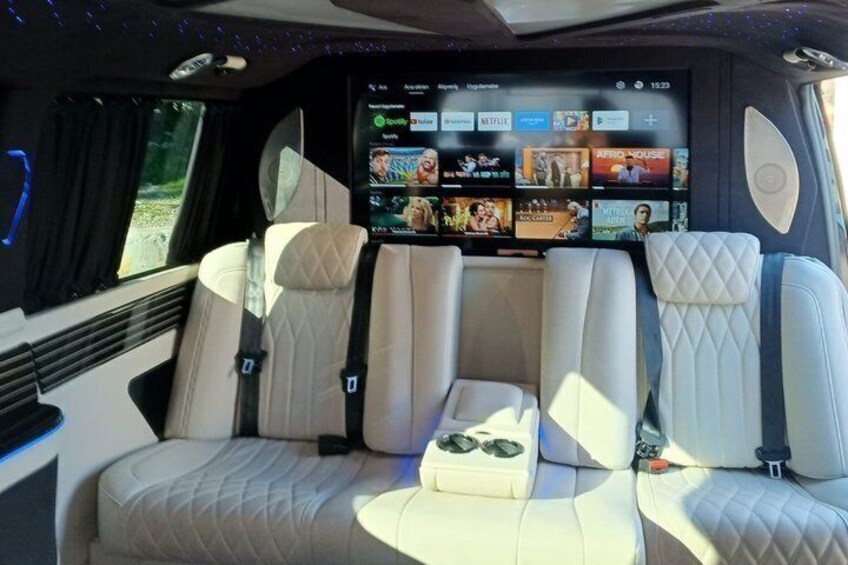 A vehicle interior with a large screen displaying various entertainment options, cushioned seating, and a control panel. 3