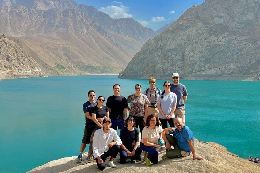 A group of people posing by a turquoise lake with mountains in the background. 2