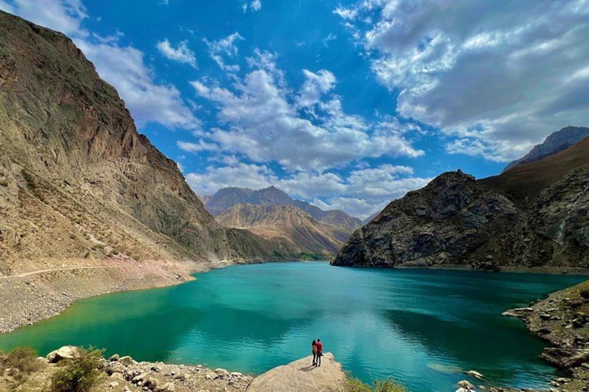Un lago turquesa rodeado de montañas, una persona de pie sobre un saliente rocoso y un cielo despejado con nubes. 1