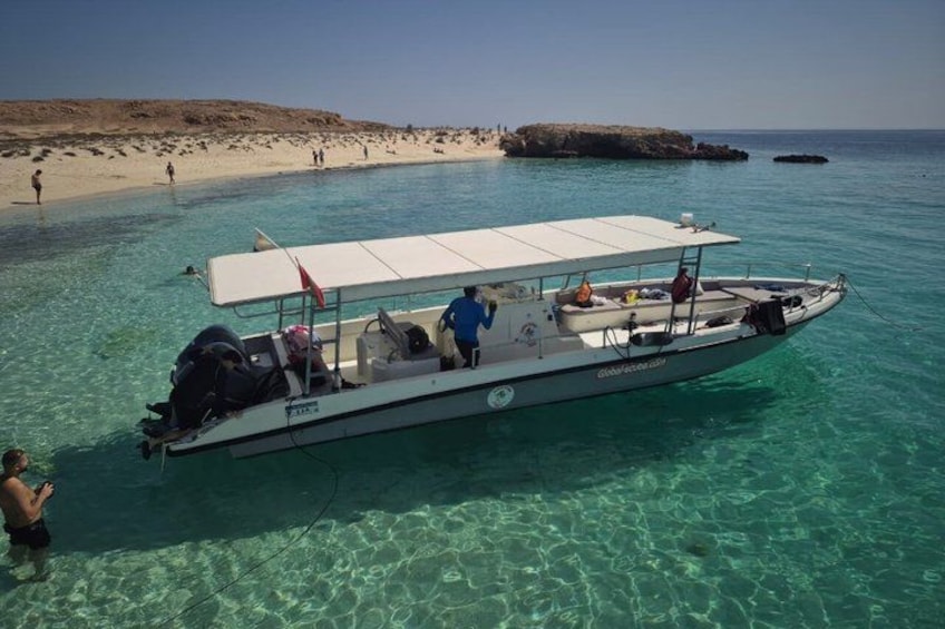 A boat on clear turquoise water with a sandy beach and rocky outcrops in the background. 1