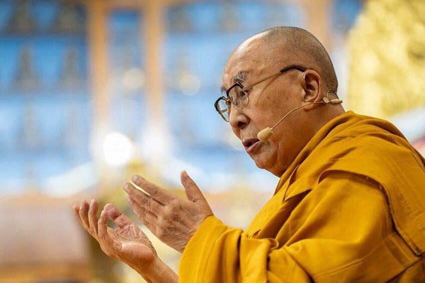 A person in a yellow robe with a microphone, hands in a prayer gesture, in an indoor setting with blurred background. 5