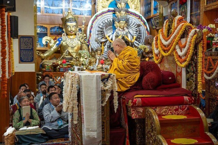 A monk in yellow robes seated on a red and gold throne, surrounded by a group of people in a temple adorned with statues and garlands. 2