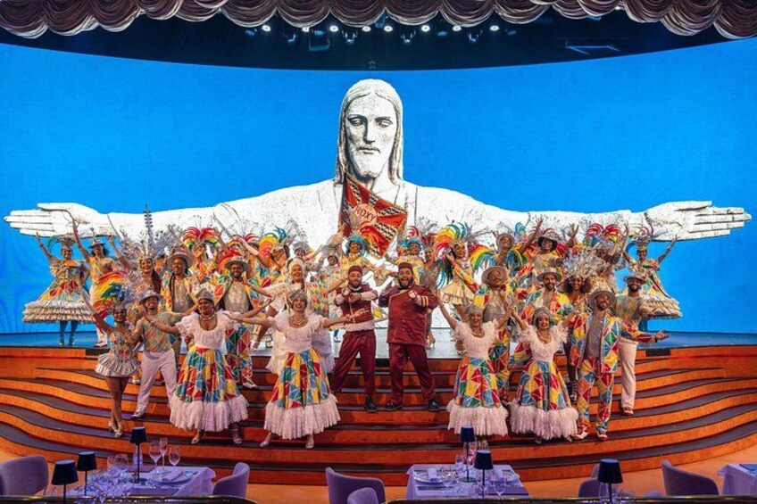 A stage with a large mural, performers in colorful costumes, and a seated figure. 2