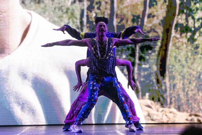 A person in a dynamic dance pose on a stage with a large screen displaying a close-up of a face in the background. 4