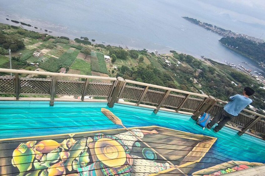 Un hombre está de pie en una terraza de madera con vista a un cuerpo de agua, y un mural colorido debajo. 4
