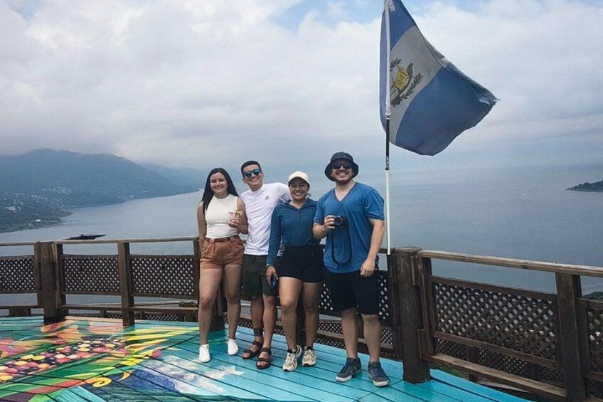 Un grupo de personas en una cubierta de madera, con una bandera, mirando un cuerpo de agua y montañas. 5