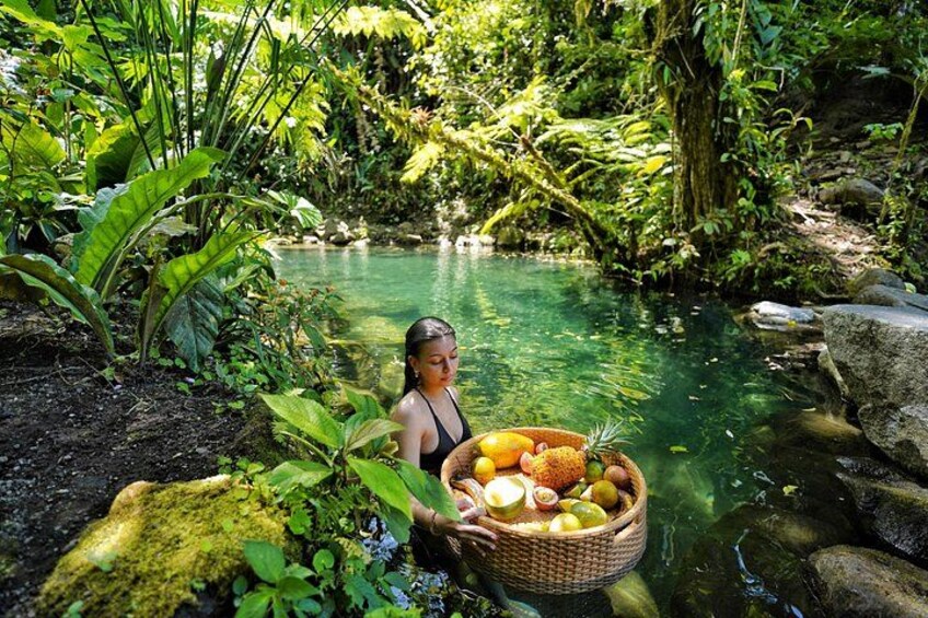 A person holding a basket of fruits by a clear water body surrounded by lush greenery. 1