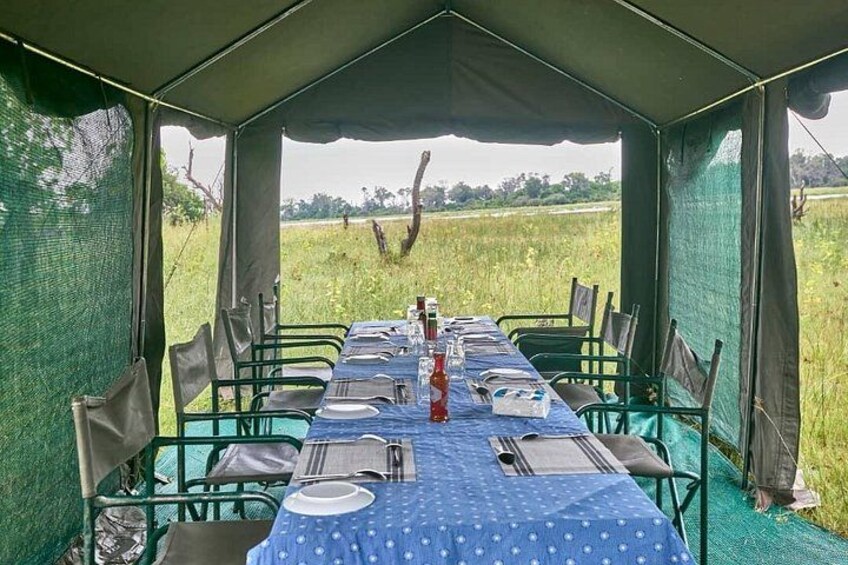 A dining table set up in a tent with chairs, a blue tablecloth, and a view of a field. 2