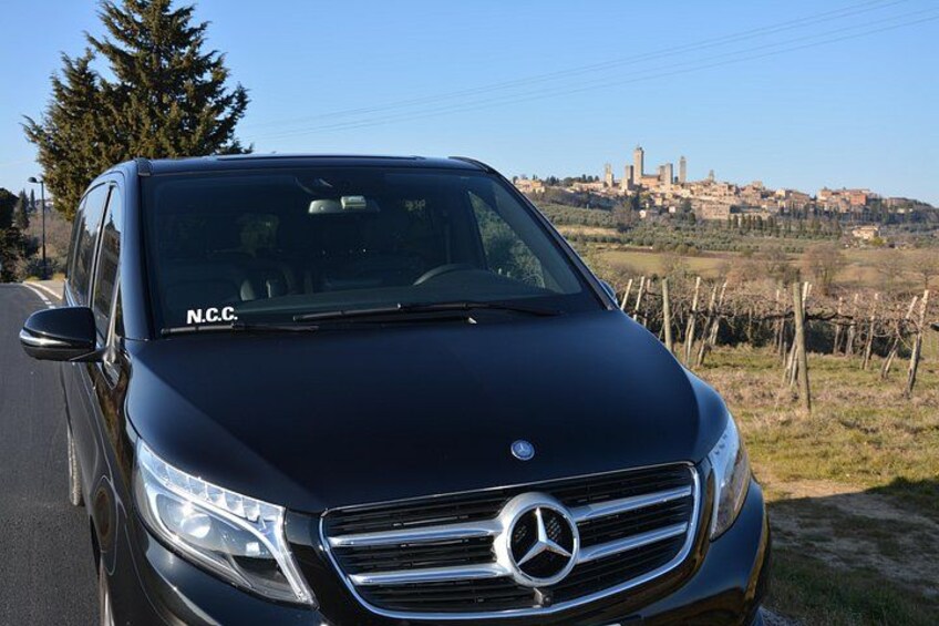 Une Mercedes-Benz noire est garée sur une route bordée d’un paysage rural, avec une colline lointaine et des bâtiments visibles à l’arrière-plan. 2