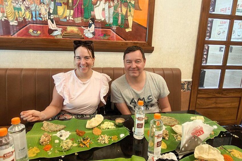 Dos personas sentadas en una mesa con una hoja de plátano, diferentes alimentos y botellas. 1