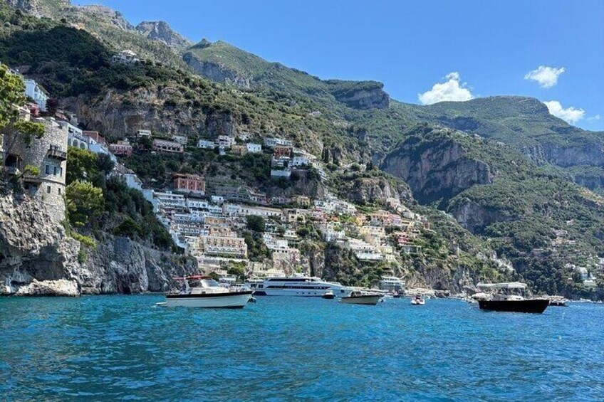 A coastal town with buildings on a hillside, boats in the water, and mountains in the background. 1