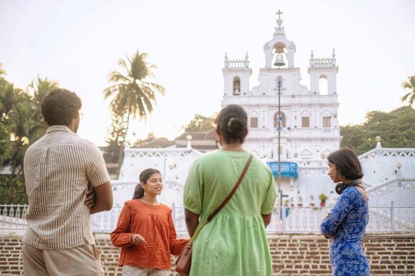 A group of people standing in front of a white colonial-style church with a bell tower, palm trees, and a brick wall in the background. 1