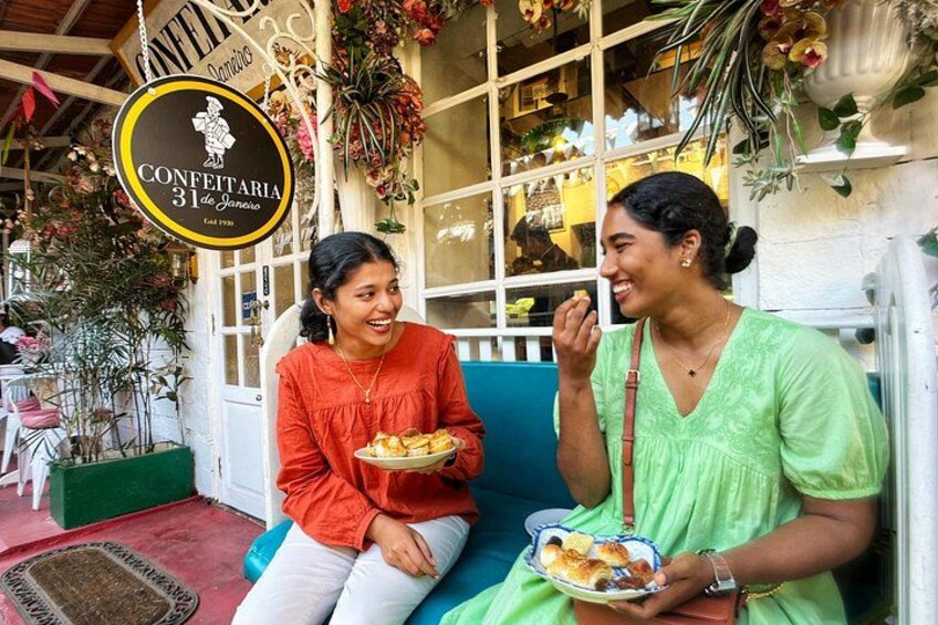 Two people sitting on a bench outside a cafe with a sign that reads 'Confetaria 31 de Enero.' 3