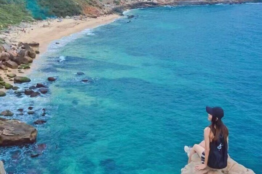 A person sitting on a rock by a clear blue sea with a sandy beach and rocks in the background. 1