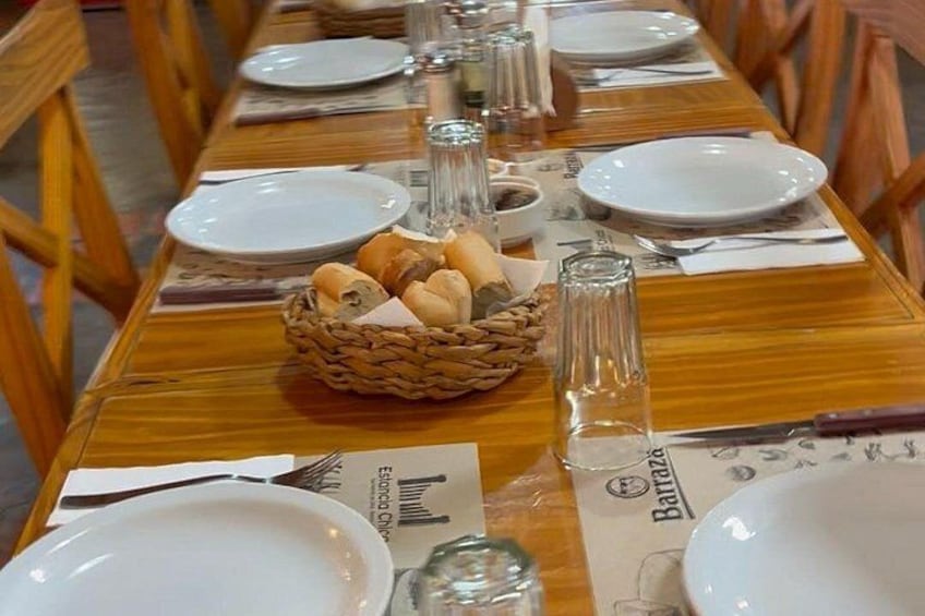 A wooden dining table set with white plates, silverware, and a basket of bread. 3