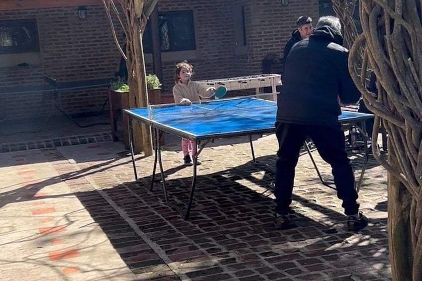 An outdoor table tennis game on a brick patio with a tree and a building in the background. 2