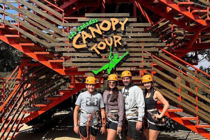 A group of people in helmets posing in front of a wooden structure with the text 'CANOPY TOUR' and a green logo. 1