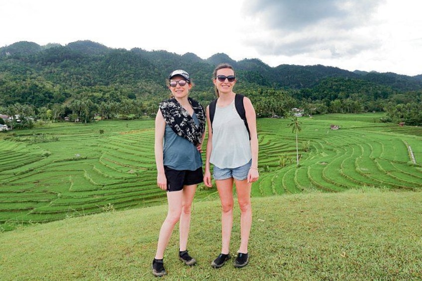 Two people standing on a grassy hill with terraced fields and mountains in the background. 3
