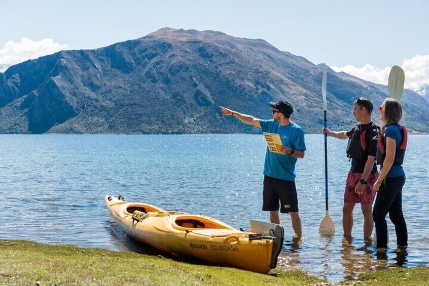 Un grupo de personas junto a un lago, con un kayak y tablas de remo. 1