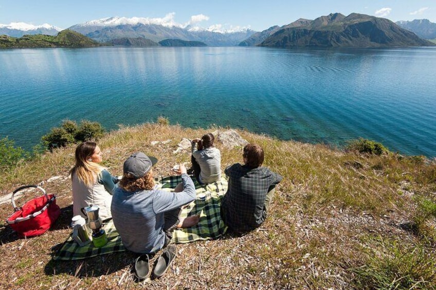 Cuatro personas sentadas sobre una manta junto a un lago, con montañas al fondo. 2