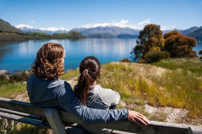 Dos personas sentadas en un banco, contemplando un lago con montañas al fondo. 5