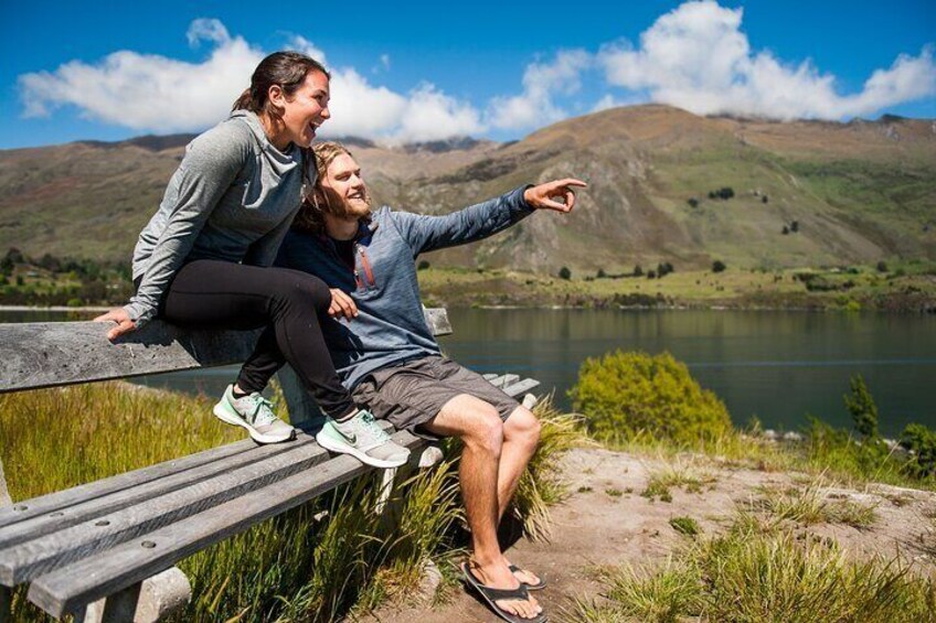 Dos personas sentadas en un banco junto a un lago, con montañas al fondo. 4