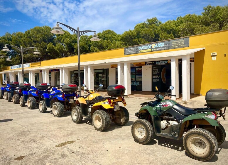 Une rangée de quads est stationnée devant un bâtiment jaune sur lequel on peut lire l’enseigne « MenorQuad.com ». 1