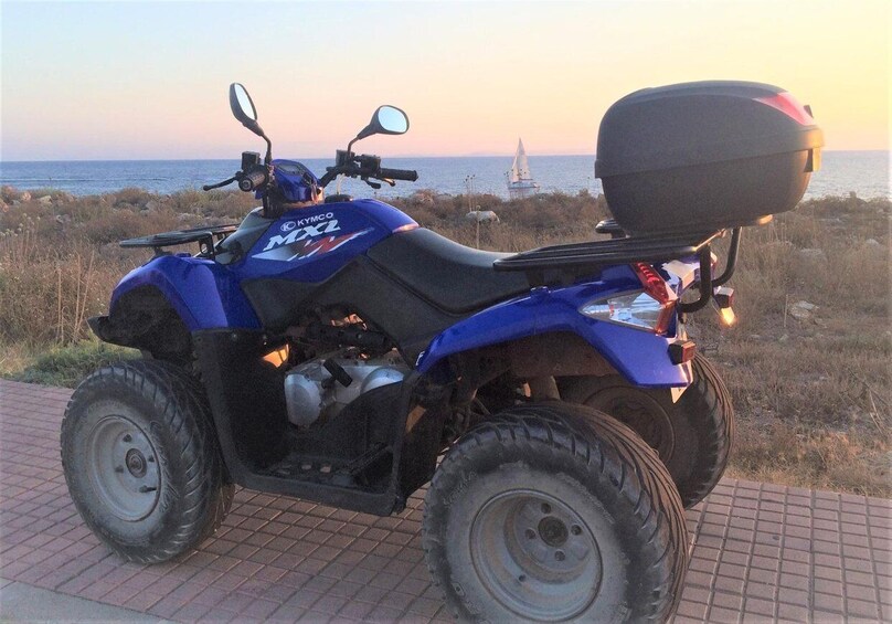 Un véhicule tout-terrain bleu de marque KYMO MX2 est garé sur une zone pavée, offrant une vue panoramique sur la mer et un voilier en arrière-plan. 3